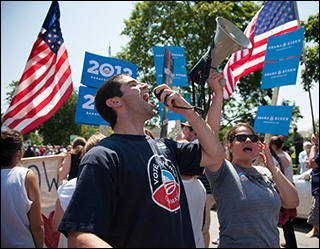 A photograph of two campaign field organizers, wearing Obama 2012 shirts. One is shouting into a megaphone and the other is cheering. The crowd behind them is holding many Obama / Biden 2012 signs and American flags.