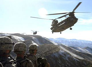 Three soldiers stand together in the mountains, waiting for two helicopters to land.