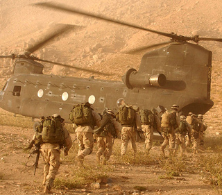 A group of U.S. soldiers run in a desert dressed in tan fatigues towards a large waiting helicopter.
