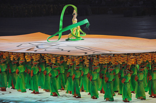 Dozens of people dressed in bright green uniforms carry poles that support a movable stage. A dancer twirling a green ribbon, stands on the stage.