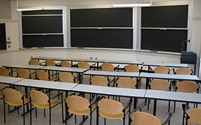 Four rows of moveable tables and wooden chairs facing a wall of blackboards.
