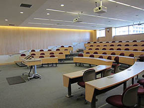 A tiered classroom with sliding chalkboards, a small table at the front for the instructor, and 2 ceiling-mounted projectors.