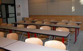 A view of a classroom from the front, with four rows of tables and chairs behind the tables.