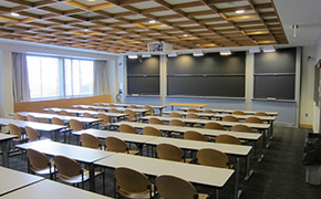 A classroom with 3 sliding chalkboards, seven rows of flat tables for students, a small table at the front for the instructor, and a ceiling-mounted projector.