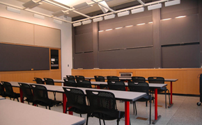 A photograph from the back of a classroom showing several rows of long tables and chairs and several blackboards up front.