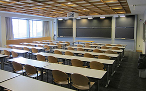 A classroom with chalkboard, several rows of flat tables for students, and a small table at the front for the instructor.