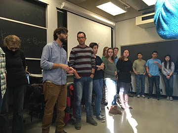 A group of people standing in a circle in a classroom before executing an Improv game.