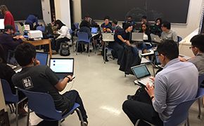 Classroom with students sitting at moveable tablet desks arranged in small groups. Students are working on laptops.